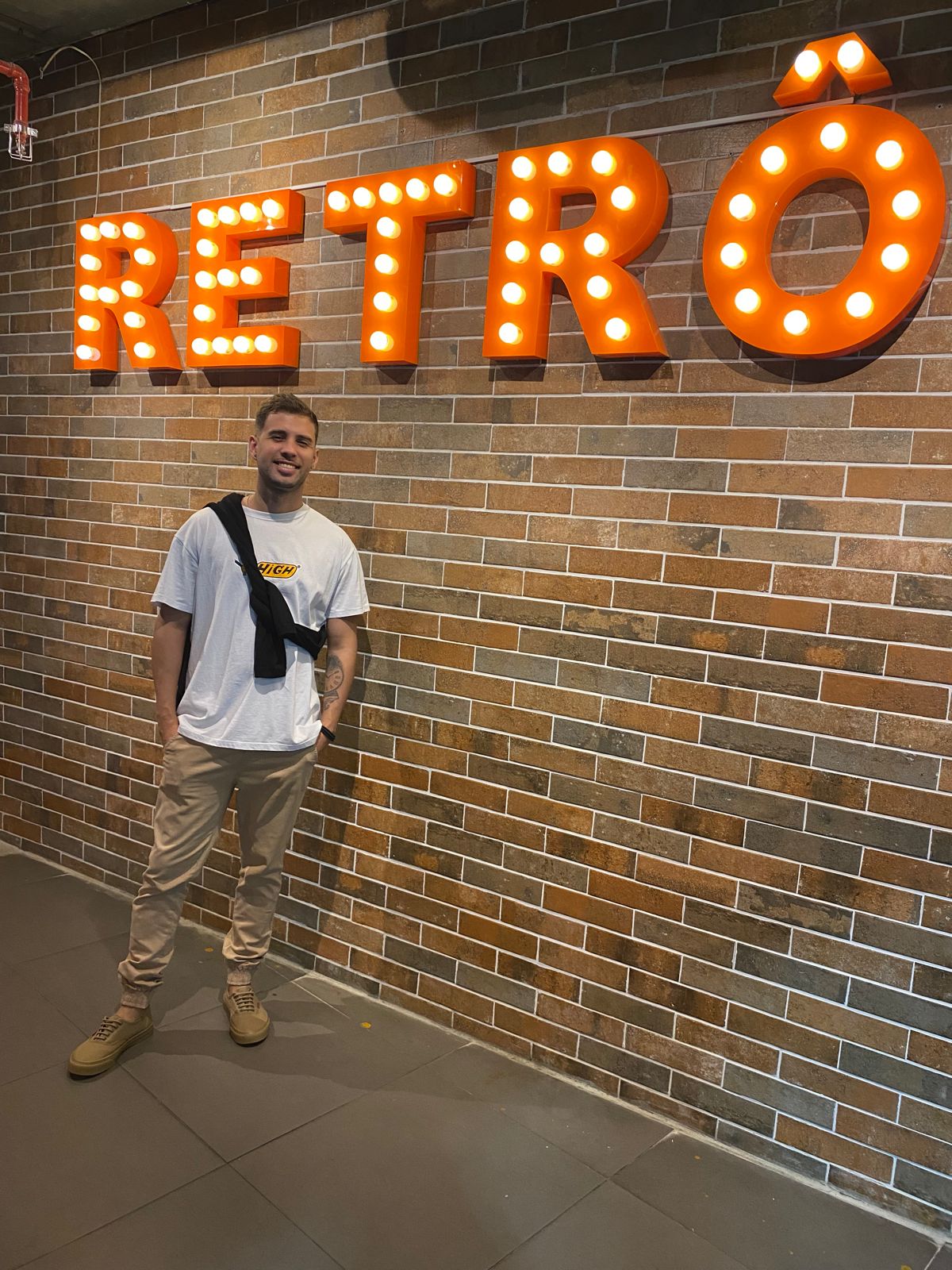 Imagem de Daniel Felipe no shopping estiloso olhando pra câmera e sorrindo, com uma logo escrito Retro atrás e uma parede de tijolos